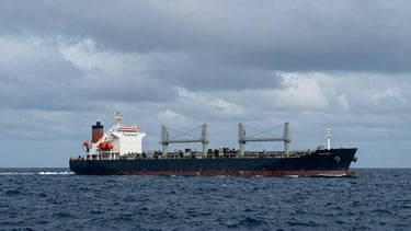 A massive cargo ship loaded with heavy machinery navigating open ocean under dramatic overcast daylight, deep steel blue tones, wide heroic framing, 35mm.