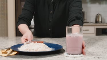 A person serving a plate of sugar, flour, jerky stick and a glass of pink milk on a kitchen counter.