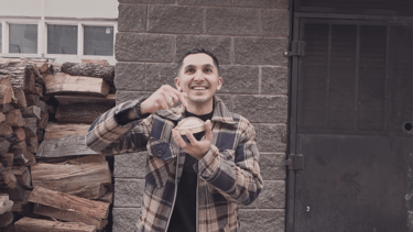 A smiling man in a plaid jacket presses a large luxury gold button against a brick wall background.