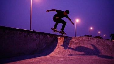 Low-angle dynamic action shot of a skateboarder performing a trick over a concrete gap at dusk, dramatic purple and magenta streetlights casting long shadows, raw concrete textures, 35mm film grain, dark purple overlay.