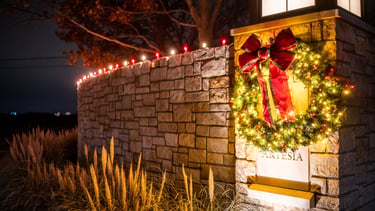 Illuminated Christmas wreath with a red bow on a stone HOA entrance at night.