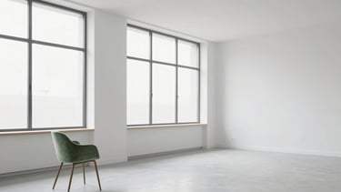 A wide-angle minimalist shot of an empty creative studio space with large windows and a single sage green chair, emphasizing white space and modern design.