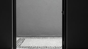 A view through a minimalist doorway showing a textured grey wall and a fragment of a monochromatic mosaic border on the floor, sharp focus, high contrast.