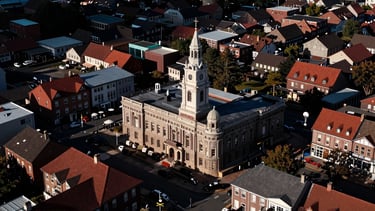 Wide environmental shot of a structured neighborhood meeting in a historic town hall, high-contrast dramatic natural light casting long shadows, 35mm.