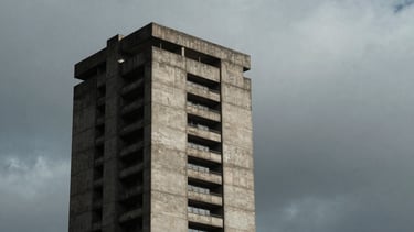 Stark architectural geometry of a concrete brutalist tower rising into an overcast, heavy gray sky, high-contrast side lighting, deep shadows, desaturated moody tones, medium-format film.