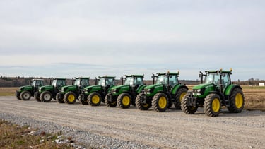 Wide panoramic landscape shot of a clean gravel equipment lot in rural Ontario under high overcast morning light, several late-model green tractors parked in a neat row, distant treeline.
