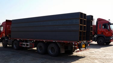 Wide-angle low shot of multiple flatbed trucks loading pre-cut structural steel beams at dawn in an industrial yard, harsh direct sunlight casting long shadows, high contrast black and red tones, 35mm lens.