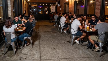 A wide-angle direct flash photograph of the concrete sidewalk outside the taproom at night, simple metal tables crowded with people, cozy chaotic arts district energy