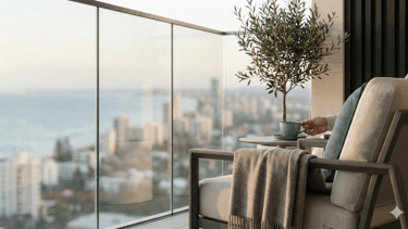Luxury modern balcony with glass railings, a lounge chair, and a scenic coastal city view at sunset.