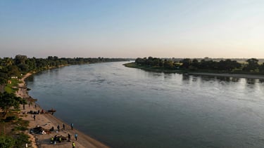 Panoramic aerial view of the Narmada river bend at golden hour, volunteer teams visible on the near bank clearing debris, distant forest on the far bank, wide cinematic frame with the river occupying the center third and sky filling the upper half