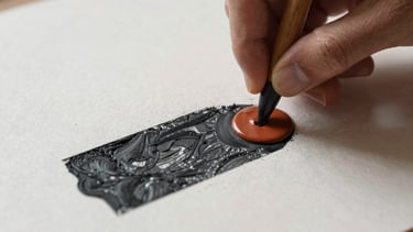 A detailed close-up of a hand rolling ink onto a carving, showcasing the texture of the paper and the rich charcoal and terracotta pigments.