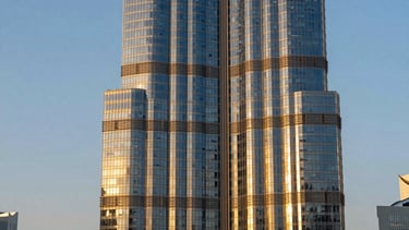 A high-end, professional architectural photograph of a luxury skyscraper in Business Bay, Dubai, UAE, during the golden hour. The building's reflective glass facade mirrors a warm sunset, and the sophisticated urban landscape conveys international business excellence and prestige. The color palette features deep blues and elegant gold reflections.