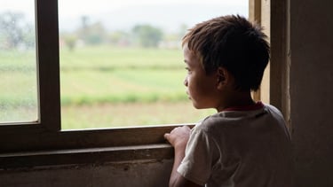 A Myanmar child sitting quietly by a window, looking out at a green landscape, soft natural daylight, shallow depth of field, warm editorial photography.