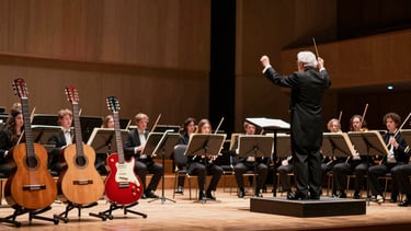 Wide panoramic view of a dramatic concert stage split in lighting. On the left third, under soft warm spotlights, a classical guitar, an acoustic guitar, and a bold red Paul Reed Smith electric guitar sit on wooden stands. On the right two-thirds, the empty chairs and polished wood instruments of a small chamber orchestra are visible, with a white-haired conductor in a tuxedo standing with his back to the audience, baton raised high in the air.