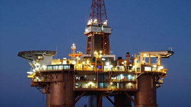 A wide-angle professional photograph of a modern offshore oil drilling platform at dusk. The scene is illuminated by warm bronze and gold safety lights contrasting with a deep navy blue sky and dark ocean waves. International / Global setting, depicting powerful engineering and modern efficiency.