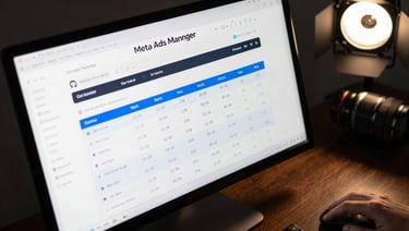 Tight shot of a monitor displaying a Meta Ads Manager campaign dashboard with cost-per-lead metrics and ROAS columns highlighted in blue, dark desk surface, sharp directional studio light from upper right, mouse hand partially visible