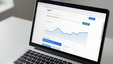 Close-up overhead of a laptop screen displaying a website analytics dashboard with organic search traffic curves, clean white desk surface, cool studio daylight from the left, keyboard partially visible at bottom edge