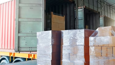 Wrapped boxes on pallets being loaded into a shipping container at a warehouse loading dock.