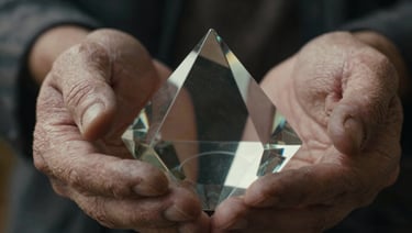 High-contrast wide anamorphic cinematic still, extreme close-up of weathered hands holding a delicate vintage glass prism, moody tungsten light leak.