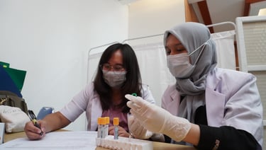 Dua tenaga laboratorium wanita mengenakan masker sedang mencatat dan memeriksa sampel darah