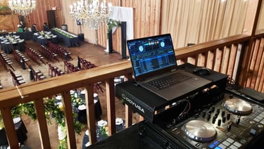 A DJ booth setup on the balcony of an event venue overlooking a wedding ceremony space.