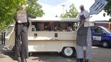 Street performers on stilts dressed as chefs posing with a vintage Citroen food truck at an outdoor event.