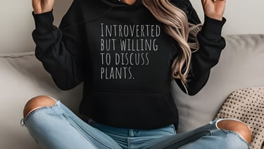 Woman in a black “Introverted but willing to discuss plants” hoodie sitting cross-legged indoors.