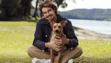 Rafael Vitti sorri, sentado na grama, abraçando um cachorro viralata caramelo.