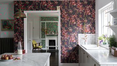 Modern kitchen with red floral wallpaper, marble countertops, and a view into a cozy living room.