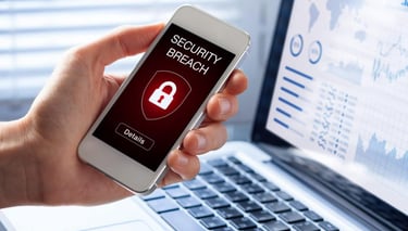 Hand holding a smartphone displaying a security breach alert next to a laptop with data charts.