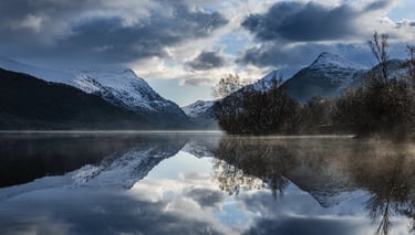 Snowy mountains reflecting in a calm, misty lake under a dramatic cloudy sky.