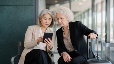 Two professional senior businesswomen looking at a digital tablet while waiting at an airport terminal.