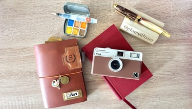 Flat lay of a leather traveler's notebook, Kodak film camera, and watercolor palette on a wooden desk.
