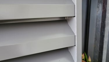 Close-up of grey exterior window shutters with fixed horizontal louvers for sun protection and ventilation.