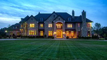 Luxury stone mansion with glowing windows and a manicured green lawn at dusk.