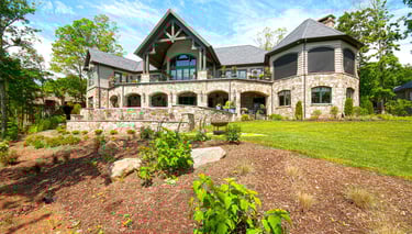 Luxury mountain estate with stone exterior, multi-level terraces, and lush landscaping under a blue sky.
