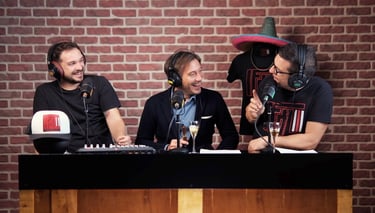 Three men recording a podcast with microphones and headphones against a red brick wall background.
