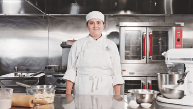 A professional female chef in a white uniform standing in a commercial kitchen with baking equipment.