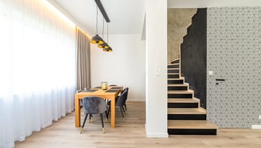 Jadalnia z drewnianym stołem i krzesłami, wiszące lampy, obok schody z czarną ścianą, mikrocement.