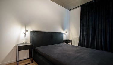 Minimalistyczna sypialnia z czarnym łóżkiem, dwiema lampkami nocnymi i zasłonami.