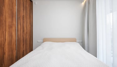 Łóżko z białą narzutą, drewniana szafa po lewej, okno z zasłonami po prawej.