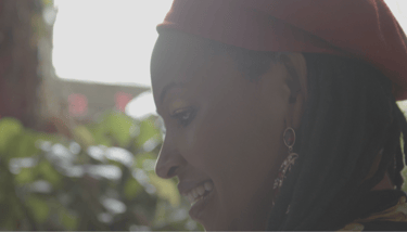 A person wearing a beanie with medium brown skin and black dreads smiling and looking down.