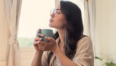 A thoughtful woman with glasses holding a coffee mug while looking out a window.