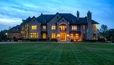 Luxury stone mansion with glowing windows and a manicured green lawn at dusk.