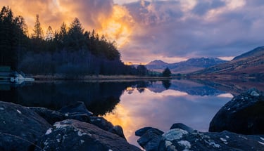 Scenic sunset reflection over a calm mountain lake with pine forest and rocky shoreline.