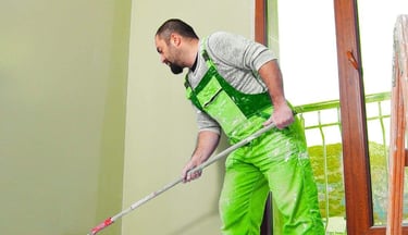 Person painting an interior wall with a roller near a window.