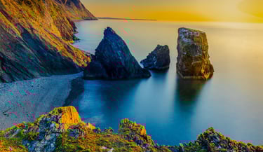 Golden hour sunset over rugged ocean sea stacks and steep coastal cliffs at a pebble beach.