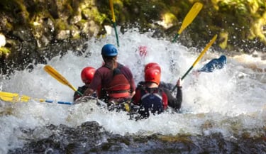 A group of people wearing helmets white water rafting through splashing river rapids with yellow paddles.