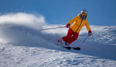 A skier in a yellow jacket and red pants carves through fresh powder on a sunny mountain slope.