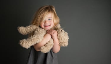 une petite fille blonde souriante faisant un calin / tenant un ours en peluche sur fond noir